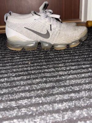Nike Air Vapormax grå sneakers - Snygga Nike Air Vapormax sneakers i ljusgrått med stickad ovandel och genomskinlig sula. Skorna har klassisk Nike-logga i svart på sidan och vita skosnören. Ovandelen är i mesh/strumpmaterial för extra komfort och stil.