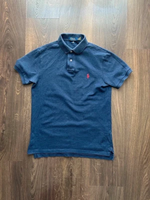 Marinblå pikétröja från Polo Ralph Lauren - Klassisk marinblå pikétröja från Polo Ralph Lauren med röd broderad logga på bröstet. Tröjan har korta ärmar, krage och knappar framtill. Perfekt till en avslappnad men ändå stilren look. Riktigt bra skick 