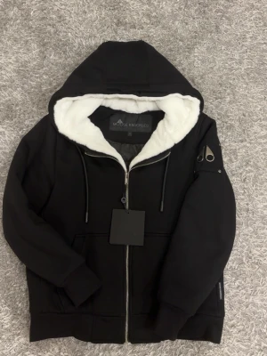 Moose Knuckles bunny S - Snygg svart hoodie från Moose Knuckles med dragkedja, vit fluffig foder på insidan och huva. Hoodien har fickor framtill, ribbade muddar och en metallpatch på ärmen. Perfekt för dig som vill ha en stilren och bekväm look.