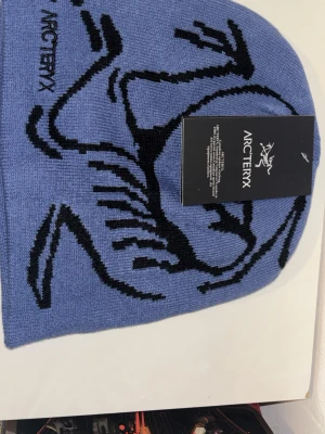 Blå mössa från Arc'teryx - Snygg blå mössa från Arc'teryx med svart grafiskt mönster och logga på sidan. Tillverkad i mjukt stickat material som sitter skönt på huvudet. Perfekt för dig som gillar outdoor och streetwear-vibe.