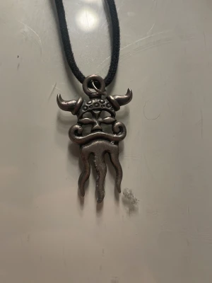 Halsband med metallhänge i vikingastil - Unikt halsband med svart snöre och ett coolt metallhänge som liknar en vikingamask med horn och vågiga detaljer. Hänget har en rå, antik look och passar perfekt för dig som gillar mytologi och statement-smycken.