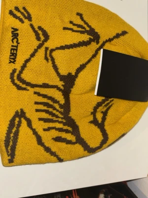Gul mössa från Arc'teryx - Snygg gul mössa från Arc'teryx med svart grafiskt mönster och logga på sidan. Mössan är stickad i mjukt material och har en avslappnad passform som passar perfekt till kalla dagar. Perfekt accessoar för dig som vill sticka ut.