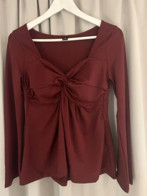 Vinröd långärmad topp med twist - Snygg vinröd topp med lång ärm och twistad detalj framtill som ger en cool v-ringad look. Tillverkad i mjukt stretchigt material som sitter skönt på kroppen. Perfekt att styla med jeans eller kjol för en trendig vibe.