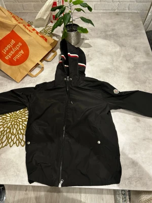 Svart Moncler vindjacka med huva - Snygg svart vindjacka från Moncler med huva och dragkedja framtill. Jackan har vita och röda ränder på huvan samt Moncler-logga på ärmen. Två fickor med tryckknappar och justerbar dragsko vid huvan. Perfekt för en sportig och stilren look.