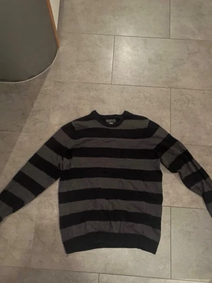 Svart och grå randig stickad tröja - Säljer en stickad tröja från Selected Homme med breda svarta och mörkgrå ränder. Tröjan har rund halsringning och ribbade muddar vid ärmslut och nederkant. Perfekt för dig som gillar en enkel men stilren look.