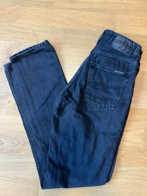 Svarta raka jeans från nudie - Snygga svarta jeans från nudie med rak passform och klassisk femficksdesign. Jeansen har en diskret logga på bakfickan och är tillverkade i ett mjukt denimtyg som känns bekvämt hela dagen. Perfekta till en avslappnad streetstyle-look.