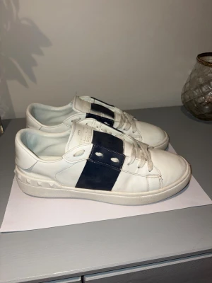 Valentino opens - Snygga vita sneakers från Valentino Garavani med bred svart rem över snörningen och ikoniska nitar på hälen. Tillverkade i läder med klassisk låg profil och vita skosnören. Perfekta för dig som gillar stilrena och exklusiva skor. Skorna har hål på sulan men är fixade