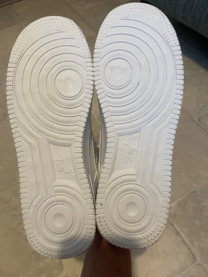 Nike Air Force 1 vita sneakers - Klassiska Nike Air Force 1 sneakers i helvitt läder med broderad Nike-logga på hälen och tjock sula. Skorna har lågt skaft, vita skosnören och en stilren design som passar till det mesta. Perfekta för dig som gillar en clean och tidlös look.