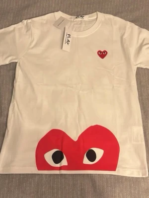 Vit t-shirt Comme des Garçons Play M - Snygg vit t-shirt från Comme des Garçons Play i bomull. Framsidan har det ikoniska röda hjärtat med ögon broderat på bröstet och ett stort tryck av hjärtat med ögon längst ner. Klassisk rund hals och korta ärmar. Perfekt statement-plagg för dig som gillar streetstyle.