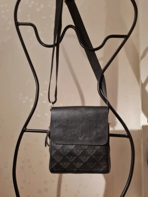 Svart axelväska från Giorgio Armani - Snygg svart axelväska från Giorgio Armani med justerbar rem. Väskan har lock med präglad logga och mönster av Armani-örnar på framsidan. Tillverkad i slätt och mönstrat konstläder som ger en exklusiv känsla. Perfekt för dig som vill ha en stilren och trendig accessoar.