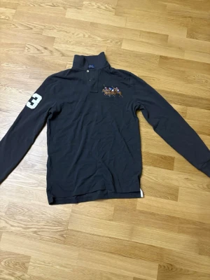 Mörkblå långärmad pikéskjorta Polo - Snygg mörkblå långärmad pikéskjorta från Polo Ralph Lauren med broderat polospel på bröstet och en vit siffra 3 på ärmen. Klassisk krage och knappar framtill. Perfekt för dig som gillar sportig och stilren look. Den har aldrig använts.  