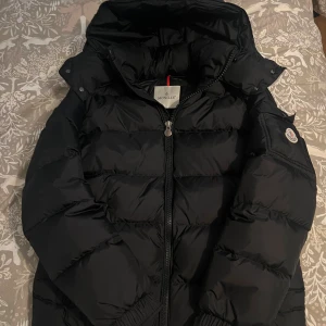 Svart Moncler pufferjacka med huva - Säljer min svarta Moncler dunjacka i mycket fint skick typ helt ny. Jackan har inbyggd NFC-scan för att verifiera äktheten. Den är extremt varm, lätt och bekväm – perfekt för kalla vinterdagar. Designen är tidlös och ger en exklusiv look som passar både till vardags och mer uppklädda tillfällen.                                            Ny pris 15000kr
