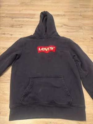 Svart hoodie från Levi's med logga - Säljer en svart hoodie från Levi's med den klassiska röda loggan på bröstet. Hoodien har en stor känguruficka framtill och en rejäl huva. Tillverkad i mjukt bomullsmaterial som är skönt att ha på sig. Perfekt för dig som gillar streetwear och enkel stil. Den har lite fläckar framme men det är inget som syns i verkligheten ❤️