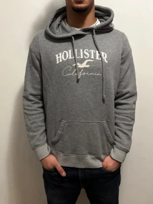 Sällsynt fodrad Grå hoodie från Hollister - Snygg grå hoodie från Hollister med vit broderad logga och texten 'California' på bröstet. Hoodien har huva med dragsko, magficka och mjukt fleecefoder på insidan. Perfekt för en avslappnad och trendig stil.