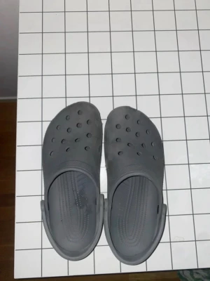 Gråa Crocs tofflor - Säljer ett par klassiska gråa Crocs tofflor i plast med ventilerande hål på ovansidan och justerbar hälrem. Perfekta för chill dagar hemma eller när du vill ha något bekvämt på fötterna. Lätta att rengöra och superbekväma.