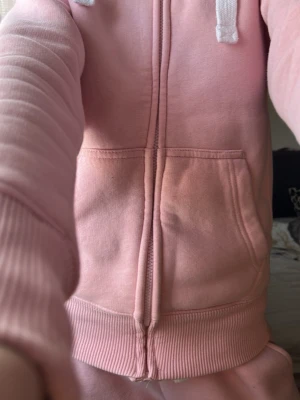 Ljusrosa russemerch hoodie - Snygg ljusrosa hoodie med dragkedja framtill och vita snören i huvan. Hoodien har känguruficka och ribbade muddar vid ärmslut och nederkant. Perfekt för en avslappnad och chill stil. Tillverkad i mjukt bomullsmaterial.