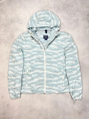 Ljusgrå mönstrad vindjacka från Woolrich - Snygg vindjacka från Woolrich i ljusgrått med vitt zebramönster. Jackan har huva, dragkedja framtill och två sidofickor. Perfekt för blåsiga dagar och enkel att matcha med streetwear. Tillverkad i lätt och smidigt material.