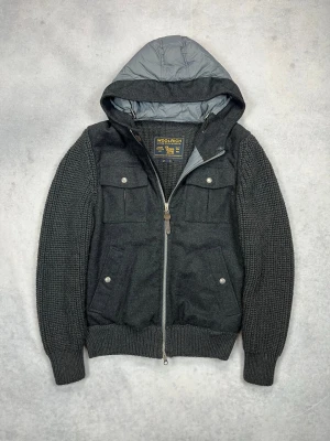 Mörkgrå stickad hoodiejacka från Woolrich - Snygg mörkgrå stickad hoodiejacka med dragkedja framtill och fyra fickor med knappstängning. Jackan har ribbade muddar, huva och en cool mix av stickat material och tygdetaljer. Perfekt för dig som vill ha en casual men ändå stilren look.