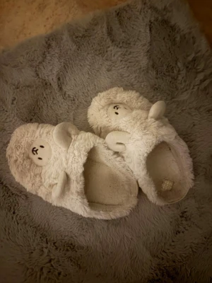 Vita fluffiga tofflor med nallebjörn - Supersöta vita tofflor i mjukt, fluffigt material med nallebjörnsansikte och öron på ovansidan. Perfekta för att hålla fötterna varma hemma och ger en gullig vibe till din outfit. Tofflorna har öppen häl och är riktigt mysiga att glida runt i.