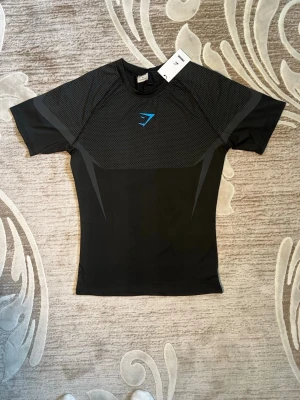 Svart träningströja från Gymshark - Snygg svart träningströja från Gymshark med korta ärmar och åtsittande passform. Tröjan har gråa detaljer och ett blått Gymshark-logo på bröstet. Tillverkad i ett stretchigt och ventilerande material som passar perfekt för gym och sport.