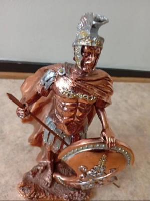 Romersk krigarstaty i bronsfärg - Detaljerad staty föreställande en romersk krigare med hjälm, spjut och sköld. Statyn är i metall-look med brons- och silverfärgade detaljer samt dekorativt mönster på skölden. Perfekt som prydnad eller samlarobjekt.