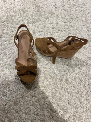 Bruna kilklackade sandaler från UGG - Snygga bruna sandaler från UGG med kilklack i kork och remmar i mocka. Skorna har öppen tå och justerbart spänne vid ankeln. Perfekta för sommaren och ger extra höjd med sin stabila wedge-klack.