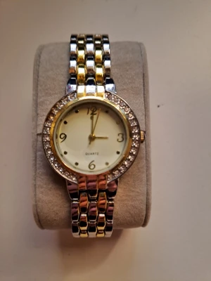 Guld- och silverfärgad klocka med stenar - Stilren klocka med rund urtavla, guldfärgade och silverfärgade länkar samt gnistrande stenar runt boetten. Urtavlan är ljus med tydliga siffror och klassisk design. Perfekt accessoar för dig som gillar en elegant look.