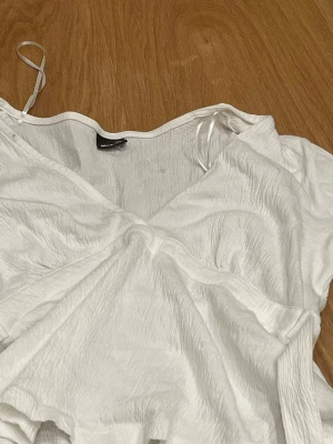 Vit omlottopp med v-ringning - Säljer en vit omlottopp från Gina Tricot med v-ringad hals och korta ärmar. Toppen har en lätt rynkad struktur och knytband i sidan för en snygg passform. Perfekt till jeans eller kjol för en fräsch look.