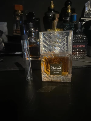 Lattafa Khamrah Eau de Parfum 10 ml - Lattafa Khamrah är en elegant parfym i en fyrkantig, kristallmönstrad glasflaska med guldbrun vätska. Flaskan har ett exklusivt utseende med detaljerad struktur och en tydlig etikett framtill. Perfekt för dig som gillar lyxiga och unika dofter.