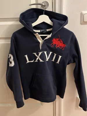 Marinblå hoodie från Polo Ralph Lauren - Passar mig 168cm lång! Snygg marinblå hoodie från Polo Ralph Lauren med stor röd logga och texten LXVII på bröstet. Stor vit siffra 3 på ena ärmen och ryggen. Klassisk känguruficka och huva. Tillverkad i mjukt bomullsmaterial, perfekt för en avslappnad och sportig stil.