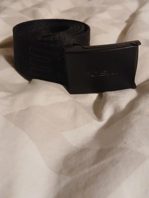 Svart tygbälte med metallspänne - Snyggt svart bälte i slitstarkt tyg med ett stilrent svart metallspänne. Perfekt för att ge en clean look till jeans eller cargo. Spännet har en enkel design och bältet är justerbart för bästa passform.