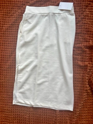 Vit pennkjol från Adoore - Stilren vit pennkjol från Adoore med hög midja och rak passform. Kjolen är knälång och har en enkel, minimalistisk design utan synliga detaljer. Perfekt att matcha med både sneakers och klackar.