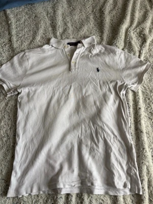 Vit pikétröja från Polo Ralph Lauren - Klassisk vit pikétröja från Polo Ralph Lauren med broderad logga på bröstet. Tröjan har korta ärmar, krage och knappar vid halsen. Tillverkad i mjuk bomull som känns skön mot huden.