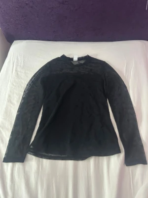 Svart mesh topp med stjärnor - Snygg svart mesh topp med lång ärm och diskreta stjärnor på ärmarna. Toppen är genomskinlig och har en tight passform, perfekt att styla lager på lager. Materialet är tunt och stretchigt syntet.