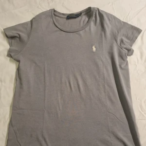 Grå t-shirt från Polo Ralph Lauren - Snygg grå t-shirt från Polo Ralph Lauren med klassisk rund halsringning och broderad vit logga på bröstet. T-shirten är i mjuk bomull och har en enkel, stilren design som passar till det mesta. 2 småsår prickar enligt sista bilden.