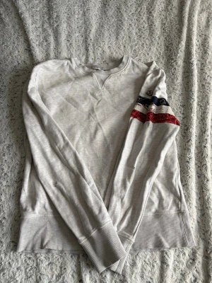 Ljusgrå sweatshirt från Moncler - Ljusgrå sweatshirt från Moncler med rund halsringning och ribbade muddar. På ena ärmen finns ett färgglatt mönster i rött, blått och vitt samt Moncler-logga. Tröjan har en avslappnad passform och är tillverkad i mjukt bomullsmaterial.