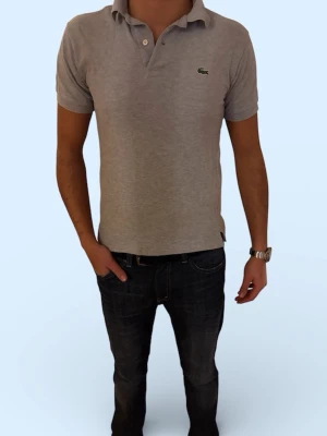 Grå pikétröja från Lacoste - Klassisk grå pikétröja från Lacoste med korta ärmar och den ikoniska gröna krokodilloggan på bröstet. Tröjan har krage och knappar vid halsen. Perfekt till jeans eller shorts för en clean och enkel stil.