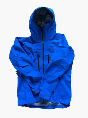 Blå vindjacka från Arc'teryx - Snygg blå vindjacka från Arc'teryx med huva och två vertikala bröstfickor med dragkedja. Jackan har svart logga broderad på bröstet och vattentäta dragkedjor. Tillverkad i slitstarkt, lätt material som står emot vind och regn.