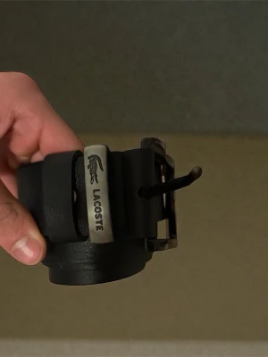 Svart skärp från Lacoste i skinn - Snyggt svart bälte från Lacoste i äkta skinn med metallspänne och logotypdetalj. Klassisk design som passar till jeans eller byxor. Perfekt accessoar för att lyfta din outfit.