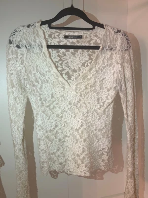Vit spetsblus från Gina Tricot - Säljer en vit långärmad blus från Gina Tricot i genombruten spets med v-ringning. Blusen har ett romantiskt blommönster och är figurnära i modellen. Perfekt att styla med jeans eller kjol för en trendig look.