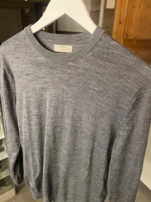 Grå merinoulltröja från MNG - Säljer en stilren grå stickad tröja från MNG i mjuk merinoull. Tröjan har rund halsringning, långa ärmar och ribbade muddar vid ärmslut och nederkant. Perfekt för lager på lager och enkel att matcha med olika outfits. Finns liten defekt på bild 2 som knappt syns.