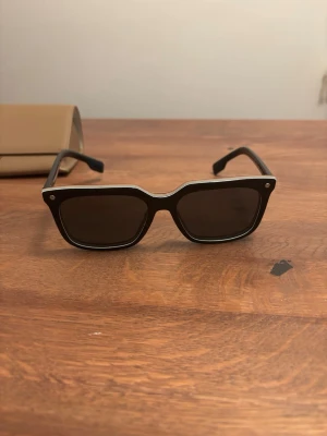 Svarta solglasögon från Burberry - Snygga svarta solglasögon från Burberry med rektangulära bågar och mörka glas. Klassisk design med diskreta silvriga detaljer i hörnen och logga på skalmen. Perfekta för att lyfta din stil under soliga dagar. Burberry solglasögon Modellen heter 4337 3798/87 56-17 Använda 3-4 gånger Ordpris: 2300kr
