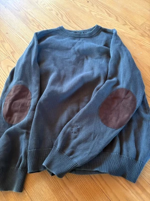Mörkgrön stickad tröja Human Scales - Snygg mörkblå stickad tröja från Human Scales med bruna mockadetaljer på armbågarna. Tröjan har rund halsringning, ribbade muddar och är tillverkad i mjukt material. Perfekt för dig som gillar klassisk men trendig stil.