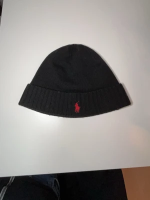 Svart mössa från Polo Ralph Lauren - Säljer en svart ribbstickad mössa från Polo Ralph Lauren med klassisk röd logga broderad framtill. Mössan har uppvikt kant och är tillverkad i mjukt material som håller dig varm under kalla dagar. Perfekt accessoar för en stilren look.