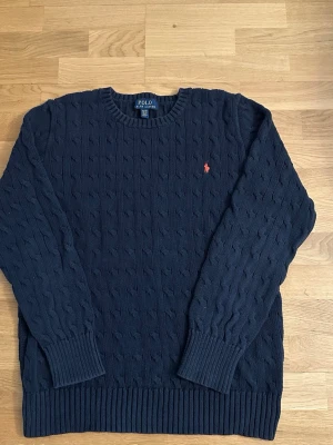 Mörkblå Ralph Lauren stickad tröja  - Kabelstickad mörkblå tröja från Polo Ralph Lauren med rund halsringning och röd broderad logga på bröstet. Tröjan har långa ärmar och ribbade muddar vid ärmslut och nederkant. Perfekt för dig som gillar klassisk och stilren look. Fraktar inom 24 timmar pris kan diskuteras 