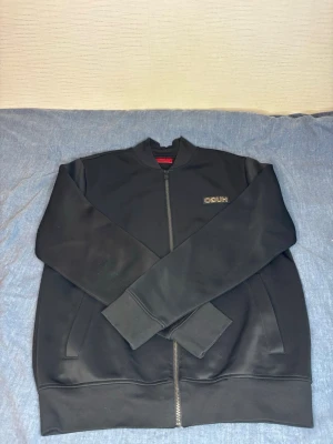 Svart zip från hugo  - Snygg svart från hugo med dragkedja framtill och diskret logga på bröstet. Tröjan har ribbade muddar vid ärmslut och nederkant samt två fickor på sidorna. Perfekt för en clean och stilren look.