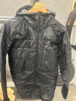 Svart vindjacka från Haglöfs - Snygg svart vindjacka från Haglöfs med huva och dragkedja framtill. Jackan har quiltad design och är tillverkad i ett glansigt, lätt material som ger en sportig look. Perfekt för kalla dagar när du vill hålla värmen utan att tumma på stilen.