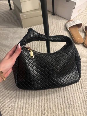 Svart flätad handväska med dragkedja - Svart handväska i flätat material med mjuk, strukturerad form. Kort handtag med knutdetalj och dragkedjestängning upptill. Rymlig invändigt trots kompakt look – funkar perfekt till vardag men även uppklätt.🖤