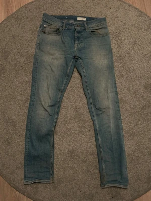 Blå slim fit jeans från tiger of sweden - Snygga blå slim fit jeans från tiger of sweden med klassisk femficksdesign och diskret slitning på låren. Jeansen har en ljus tvätt och är tillverkade i denim med en liten lagning på ena benet. Perfekta för en avslappnad och trendig look.