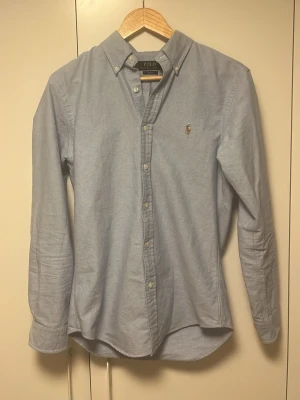 Ljusblå skjorta från Polo Ralph Lauren - Klassisk ljusblå skjorta från Polo Ralph Lauren i slim fit-modell. Skjortan har knappar framtill, button-down-krage och den ikoniska broderade loggan på bröstet. Tillverkad i mjuk bomull och passar perfekt till både jeans och chinos.